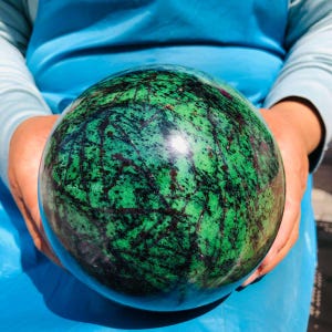 May include: A polished spherical stone featuring a vibrant green base color with intricate, dark black and deep red vein patterns throughout the surface. The sphere has a smooth, reflective finish that highlights the natural mineral inclusions.
