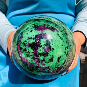 May include: A polished spherical mineral specimen featuring a vibrant mix of bright green zoisite, deep black hornblende inclusions, and striking magenta-colored ruby patches distributed across the smooth, rounded surface of the stone.