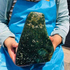May include: A large, irregularly shaped slab of dark green moss agate featuring deep forest green tones, translucent patches, and scattered white and cream-colored mineral inclusions throughout the polished, smooth surface of the stone.