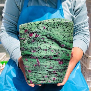 May include: A large, polished slab of zoisite mineral featuring a vibrant green base color, speckled with numerous small black hornblende inclusions and scattered, irregular patches of deep magenta-colored ruby crystals throughout the stone.