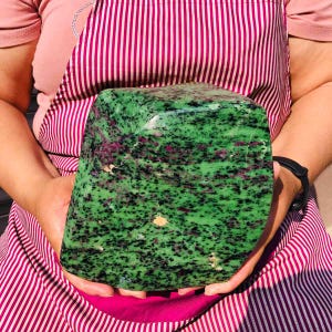 May include: A large, polished zoisite stone featuring a vibrant green base color with prominent black mineral inclusions and scattered deep magenta ruby patches throughout the surface, displaying a smooth, rounded, and irregular block-like shape.