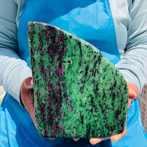 May include: A polished slab of zoisite mineral featuring a vibrant green base color with prominent streaks of deep magenta ruby and scattered black hornblende inclusions, shaped with a curved top edge and a flat base for display purposes.