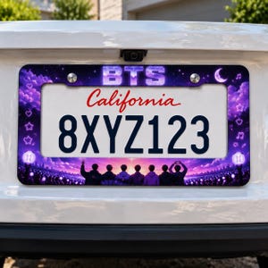 May include: A rectangular license plate frame featuring a purple galaxy-themed design with the text BTS at the top. The frame displays a concert scene with seven silhouettes, glowing light sticks, stars, musical notes, and a crescent moon.
