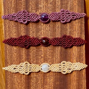 Könnte beinhalten: Drei Makramee-Armbänder mit komplizierten Knotenmustern und einem einzelnen zentralen Edelstein. Das obere Armband ist lila mit einem dunkelvioletten Stein, das mittlere ist dunkelrot mit einem dunklen Stein und das untere ist cremefarben.