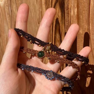 Op de afbeelding: Drie macramé armbanden met ingewikkelde gevlochten koordpatronen in zwart en bruin. Elke armband is geaccentueerd met kleine metalen kralen en een enkele centrale edelsteen, waaronder een donkergroene steen en een doorschijnende grijze steen.
