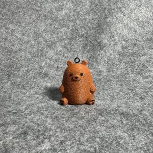 May include: A brown bear figurine with a small loop at the top for hanging. The bear has small arms and feet, and a textured surface. The bear is sitting upright.