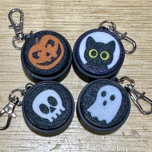 May include: Four Halloween-themed keychains with silver clasps. Each keychain features a round, black base with a glittery texture. The designs include a jack-o'-lantern, a black cat, a skull, and a ghost, all in contrasting colors.