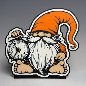 May include: A whimsical gnome-shaped clock with an orange hat and beard, holding a round clock. The clock face has black numbers and hands. The gnome is outlined in black and sits on a black base. A charming decorative item.