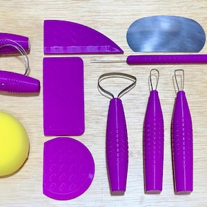 May include: A collection of pottery tools, including a wire cutter, various purple-handled tools, a metal blade, and a yellow sponge. The tools are arranged on a light-colored wooden surface, ideal for sculpting and shaping clay.