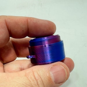 May include: A cylindrical object with a blue and purple gradient. The object has a small, protruding loop on its side. The colour transitions from blue to purple, creating a metallic sheen. The object is held in a hand.