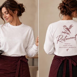 May include: A white long-sleeve shirt featuring a maroon graphic on the back with a bird illustration, the text "Restaurant", "PEOPLE GOOD FOOD BEAUTIFUL MOMENTS", and a small heart. The shirt is paired with a solid maroon waist apron.