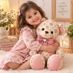 Pode incluir: Um urso de peluche em tecido cor de creme com um padrão floral repetitivo em rosa e verde. O urso tem um grande laço de tecido rosa sólido à volta do pescoço, acentuado com um pequeno pendente em forma de coração no centro e solas em tecido rosa.