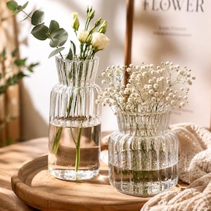 Può includere: Due vasi in vetro trasparente con una texture a coste verticali e un bordo smerlato. Il vaso più alto contiene steli di eucalipto e fiori bianchi, mentre il vaso più basso e largo contiene un mazzetto di piccoli fiori bianchi di gipsofila.