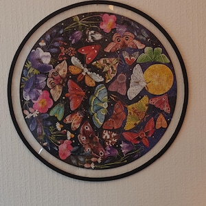 May include: A circular jigsaw puzzle framed in a thin black border, featuring a dark background filled with various colorful moths, small flowers, and a prominent yellow moon. The artwork displays a variety of moth species in shades of red, orange, blue, and brown, interspersed with botanical elements.