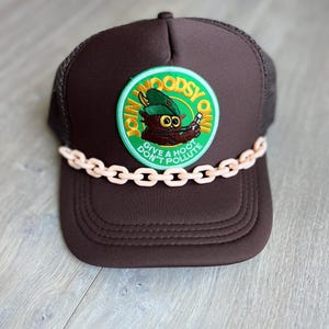 May include: A brown trucker hat featuring a mesh back and a circular embroidered patch on the front. The patch depicts a brown owl character wearing a green hat with the text JOIN WOODSY OWL GIVE A HOOT DON'T POLLUTE. A light peach plastic chain trim sits across the brim.