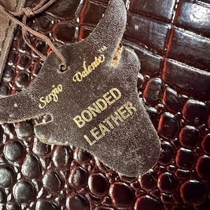 May include: A dark brown, glossy, crocodile-embossed texture surface with a bull-shaped tag attached by a brown string. The tag features the gold text Sergio Valente and BONDED LEATHER, indicating the material composition of the item.