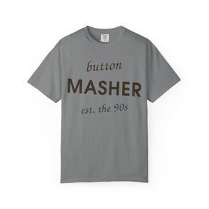 May include: A grey short-sleeved t-shirt featuring the text button MASHER est. the 90s printed in dark brown across the chest. The shirt has a crew neckline and a standard fit, with a small brand label visible on the inside of the collar area.
