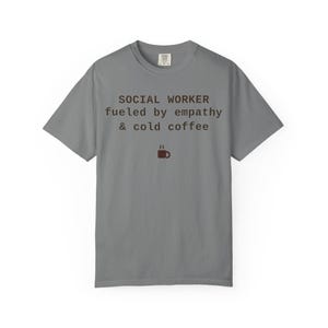 May include: A grey short-sleeved t-shirt featuring the dark brown text "SOCIAL WORKER fueled by empathy & cold coffee" centered on the chest. Below the text is a small dark brown icon of a steaming coffee mug. The shirt has a crew neckline.
