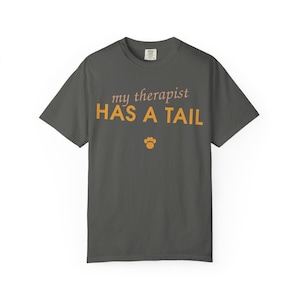 May include: A charcoal gray short-sleeve t-shirt featuring the text "my therapist" in a light pink script font above the text "HAS A TAIL" in a bold orange sans-serif font, with a small orange paw print icon centered below the text.