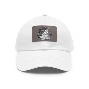 May include: A white baseball cap featuring a rectangular grey patch on the front. The patch displays a circular graphic with a landscape, a lighthouse, and the text "I LIVE IN YOUR HAPPY PLACE" and "DON'T FUCK IT UP" in black lettering.