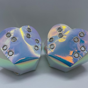 Puede incluir: Dos bolsas blancas iridiscentes en forma de corazón con ojales plateados. Las bolsas están hechas de un material brillante y holográfico.