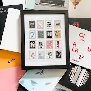 May include: A framed cross-stitch artwork featuring a grid of sixteen individual postage stamp designs. Each stamp displays unique colourful icons, symbols, and text related to the music group BTS, including titles like Wings, BE, and Butter, set against a white fabric background.