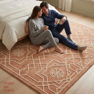 May include: A rectangular area rug featuring a terracotta-colored base with a raised, cream-colored geometric pattern consisting of interconnected lines and circular star-like motifs. The rug is textured with a soft pile and includes the text STUDIO PILE CRAFTS.