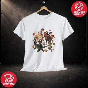 Può includere: Una t-shirt bianca a maniche corte con una grafica centrale che raffigura un Bigfoot, una creatura simile a una falena, un alieno verde e il mostro di Loch Ness che si tengono per mano attorno a un fiore, tra stelle arancioni.