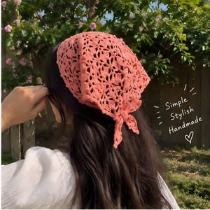 Könnte beinhalten: Ein korallenfarbenes, dreieckiges Häkelkopftuch mit einem komplizierten, durchbrochenen Blumenmuster, das hinten mit zwei kleinen Knoten gebunden ist. Der Text "Simple Stylish Handmade" mit einem Herzsymbol ist rechts zu sehen.