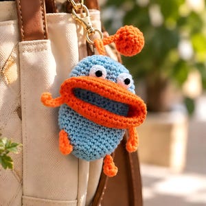 May include: A light blue crocheted creature keychain with a large orange mouth, two white and black eyes, and a small orange ball on top. The figure has small orange arms and feet and is attached to a gold-colored metal lobster clasp.