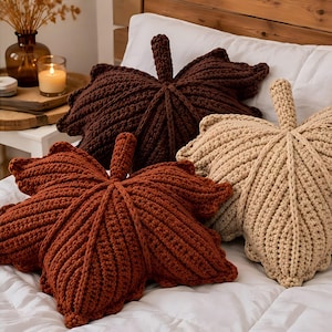 May include: Three leaf-shaped throw pillows made of chunky knit yarn. The pillows are colored in dark brown, rust orange, and beige. Each pillow features a central stem and textured, ribbed veins radiating outward to the pointed edges.
