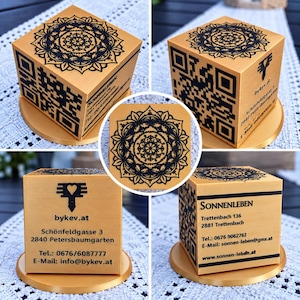 May include: A gold-colored cube featuring a black mandala design on the top surface. The sides display black QR codes, contact information for bykev.at and Sonnenleben, including addresses, phone numbers, and email addresses, all on a gold base.
