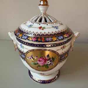 May include: A white porcelain potpourri jar with a lid, featuring a central gold oval medallion with a hand-painted floral bouquet. The jar is decorated with deep blue and gold bands, intricate floral patterns, and has two side handles.