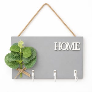 May include: A rectangular gray wall-mounted key holder featuring the white text HOME, three white metal hooks, and a decorative green eucalyptus sprig tied with a jute twine bow. The unit is suspended by a thick tan rope attached to the top.