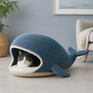 May include: A blue crocheted pet bed shaped like a whale with a wide open mouth, featuring a cream-colored interior lining and a soft white cushion inside. A grey and white cat is resting comfortably within the whale-shaped structure.