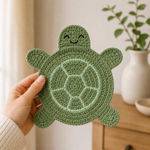 May include: A round, green crochet coaster shaped like a turtle. The coaster features a darker green outer edge and a lighter green central shell pattern with stitched segments. The turtle head at the top has a simple embroidered smiling face.