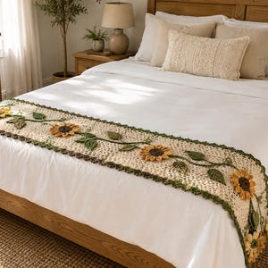 Könnte beinhalten: Ein cremefarbener, gehäkelter Bettläufer mit einem wiederkehrenden Muster aus dreidimensionalen gelben Sonnenblumen mit dunkelbraunen Mitten, grünen gehäkelten Blättern und einem dunkelgrünen, gebogten Rand auf einer weißen Tagesdecke.