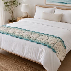 May include: A rectangular bed runner featuring a cream-colored crochet pattern of repeating seashell motifs. The runner is bordered by a scalloped edge in a vibrant teal blue yarn, creating a textured decorative accent across the white bedding.