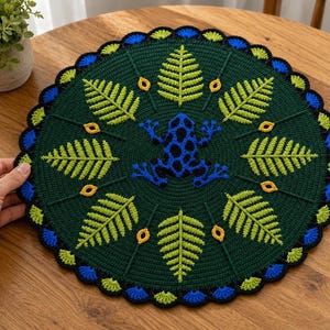 May include: A circular crochet doily featuring a dark green background with a central blue and black frog motif. The design includes radiating light green fern leaves and small yellow accents, finished with a scalloped border of blue and green.