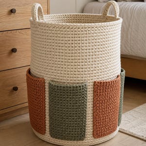 May include: A cylindrical cream-coloured crochet basket featuring two sturdy integrated handles at the top. The exterior is decorated with alternating rectangular pockets in muted sage green and terracotta orange, crafted with a textured stitch.