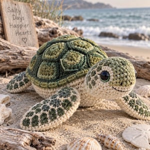 Op de afbeelding: Een gehaakt zeeschildpad knuffeltje met een donkergroen en olijfkleurig schild, een crèmekleurige onderkant en grote zwarte ogen. De schildpad ligt op het zand bij een houten bordje met de tekst Salty Days Happier Hearts en een hartje.