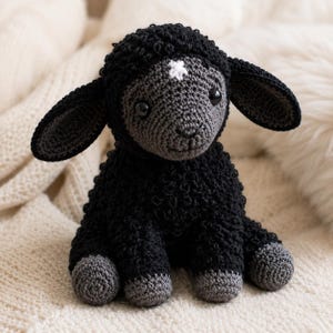 May include: A crocheted stuffed animal in the shape of a lamb. The lamb has a black textured body, dark gray face and hooves, and a small white star-shaped detail on its forehead. It features large black plastic eyes and floppy ears.