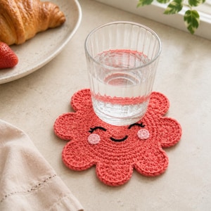 May include: A coral-colored, flower-shaped crochet coaster featuring an embroidered smiling face with black eyes, a curved mouth, and light pink circular cheeks. A clear, ribbed glass filled with water sits centered on top of the coaster.