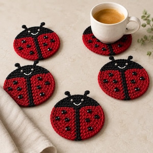 Può includere: Un set di quattro sottobicchieri rotondi all'uncinetto a forma di coccinella, caratterizzati da colori rosso e nero con pois di perline nere, sorrisi ricamati in bianco e due piccole antenne. Una tazza bianca in ceramica contiene caffè.