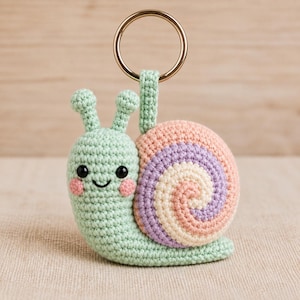 Puede incluir: Un llavero de caracol tejido a ganchillo con cuerpo verde claro, dos antenas, ojos negros y mejillas rosadas. La concha tiene un patrón en espiral con tonos pastel alternos en rosa, lavanda y crema. Incluye una argolla de metal dorado.