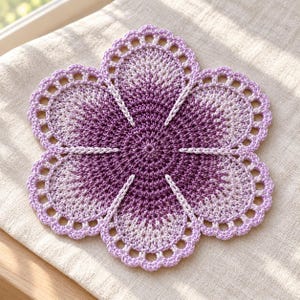 May include: A circular crochet doily featuring a flower-like shape with five rounded petals. The design transitions from a deep purple center to lighter lavender shades at the edges, finished with a delicate scalloped lace border.