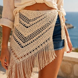 May include: A cream-colored crochet wrap skirt featuring an openwork chevron pattern, long fringe trim along the bottom edge, and side ties with decorative tassels. The garment is worn over dark blue denim shorts with a frayed hemline.