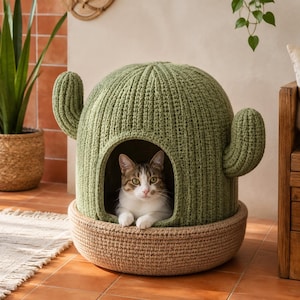 May include: A sage green crocheted cat bed shaped like a cactus with two side arms and a rounded top. The base is a tan, woven circular structure. A brown and white tabby cat is resting inside the arched opening of the cactus-shaped enclosure.