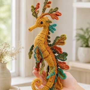 Op de afbeelding: Een gehaakt zeedraak-speeltje met een textuurrijk mosterdgeel lichaam en ingewikkelde, veelkleurige aanhangsels in tinten bosgroen, olijf en gebrand oranje. Het wezen heeft een gekrulde staart en een lange snuit met een klein zwart oogje.