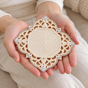 May include: A cream-colored crochet coaster with a quatrefoil shape, featuring a solid circular center and an intricate openwork border. The outer edge is accented with small, clear, reflective sequins along the silver-toned trim.